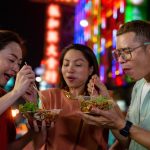 Tiga wisatawan mencicipi makanan lokal saat menjelajahi tempat makan di luar negeri pada malam hari.