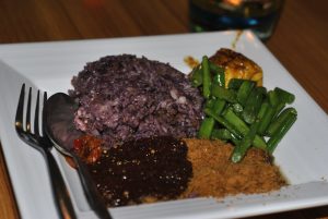 Sepiring Nasi Kalong berwarna hitam keunguan, disajikan dengan lauk seperti tumis buncis, ayam, serundeng, sambal, dan saus hitam khas