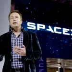 Elon Musk berbicara di atas panggung dengan logo besar SpaceX yang ditampilkan pada layar di belakangnya