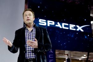 Elon Musk berbicara di atas panggung dengan logo besar SpaceX yang ditampilkan pada layar di belakangnya